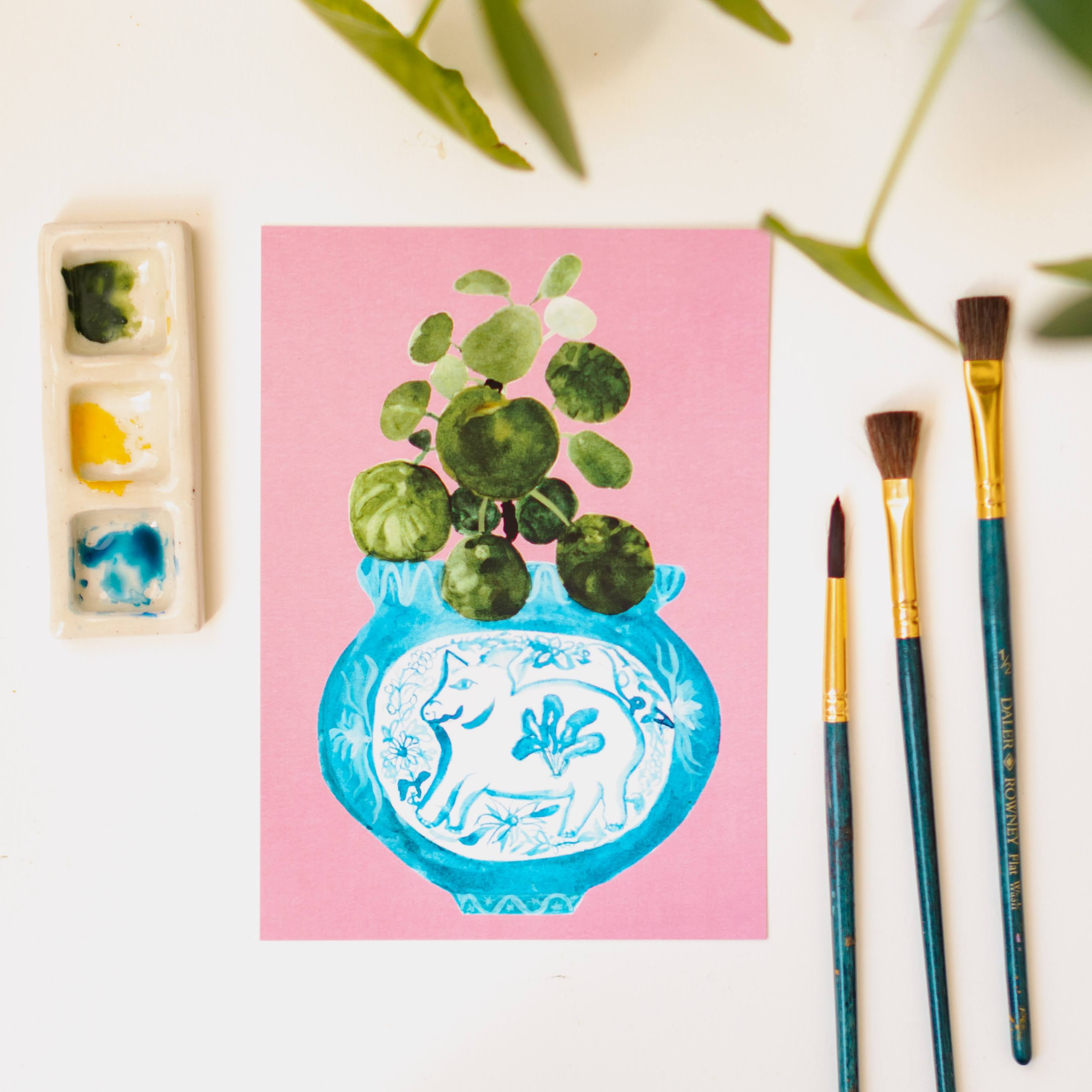 A print of a pilea plant in a blue and white porcelain vase, decorated with a pig.