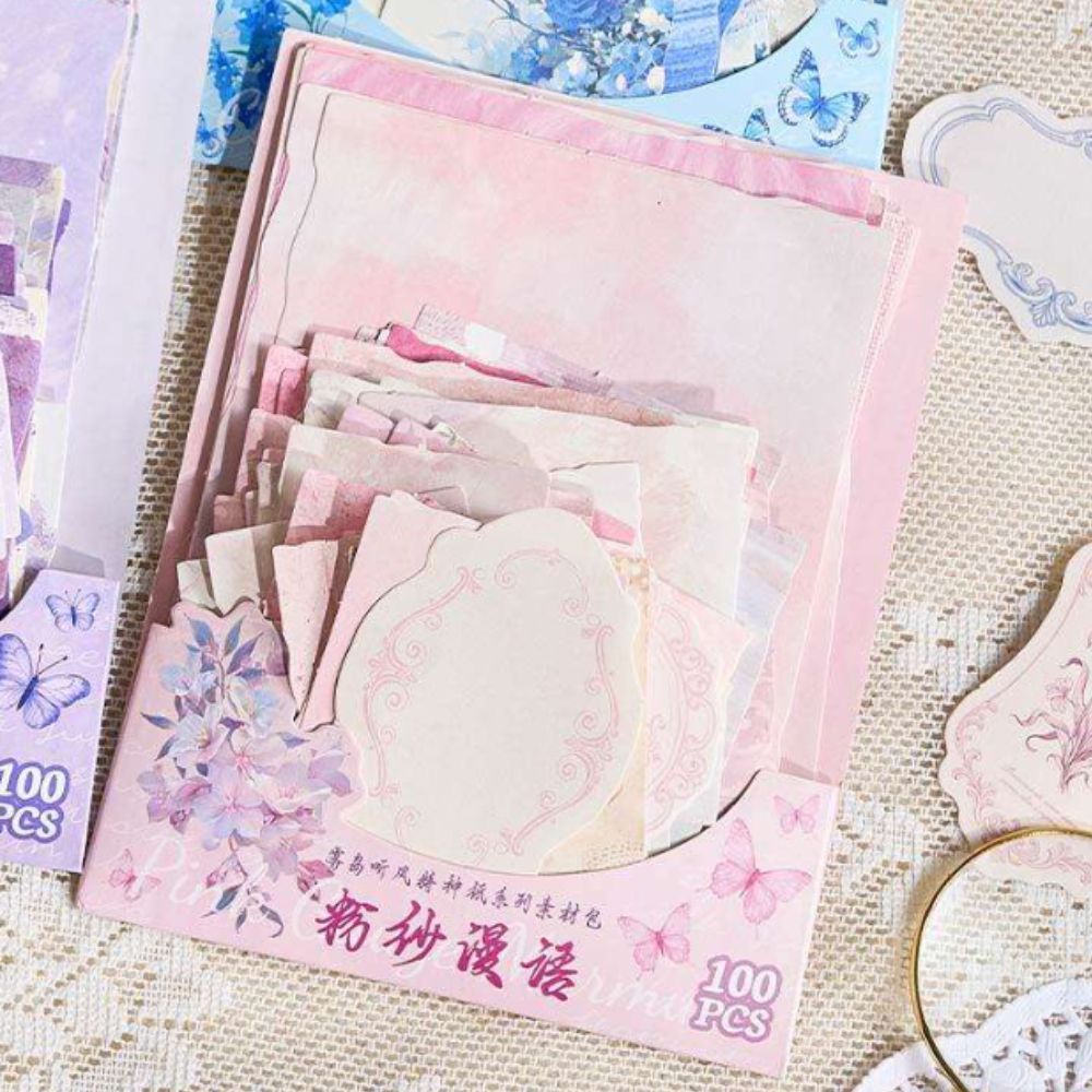 A pink decorative paper pack labeled '100 PCS' with layered pastel cardstock sheets and floral butterfly graphics, photographed on a textured beige fabric surface.