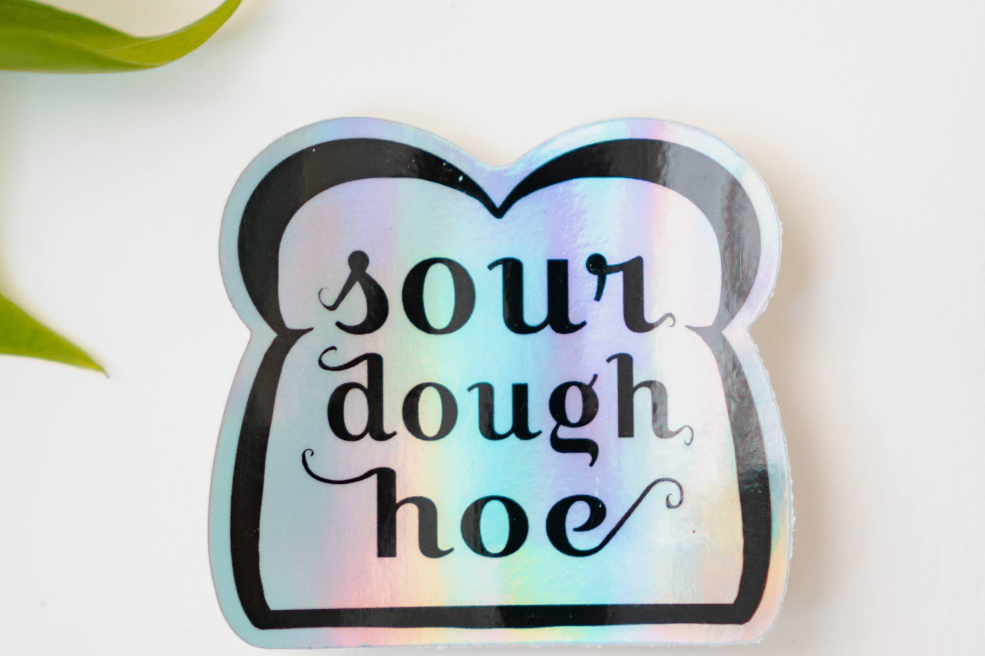 A holographic sticker featuring line art of a slice of bread and the words "sourdough hoe" written in the center.