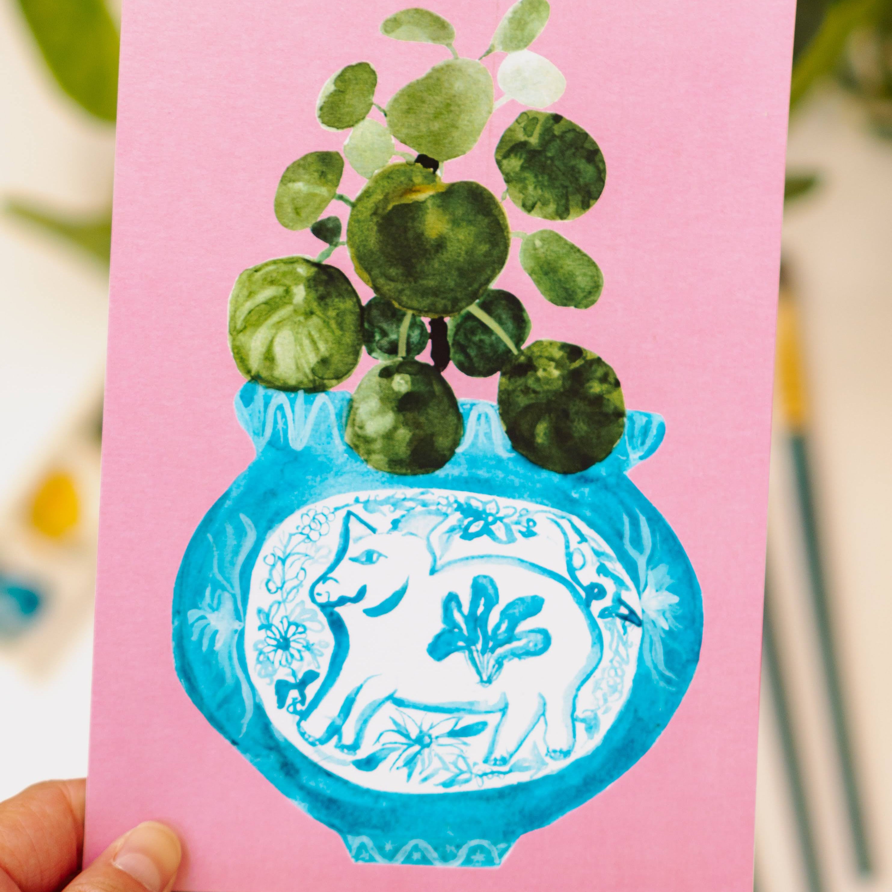 A print of a pilea plant in a blue and white porcelain vase, decorated with a pig.