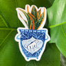 A sticker of a blue and white vase with a dog on it. The vase contains a snake plant.