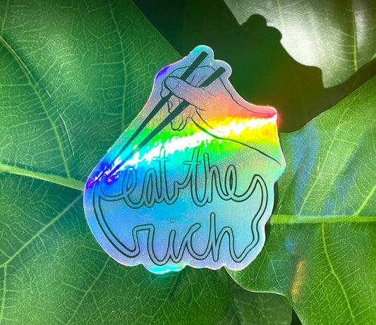 A holographic sticker featuring someone with a pair of chopsticks picking up a noodle which spells, "eat the rich."