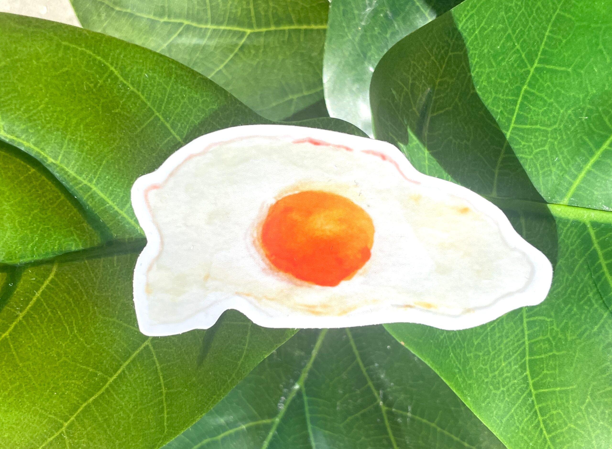 A narrow, white sticker of a fried, sunny side up egg with a rich yellow yolk and crispy edges.