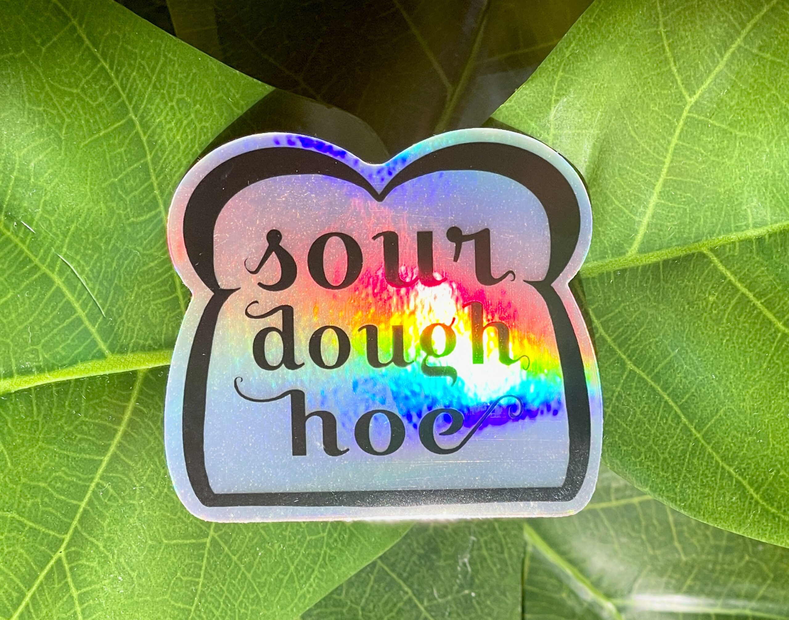 A holographic sticker featuring line art of a slice of bread and the words "sourdough hoe" written in the center.