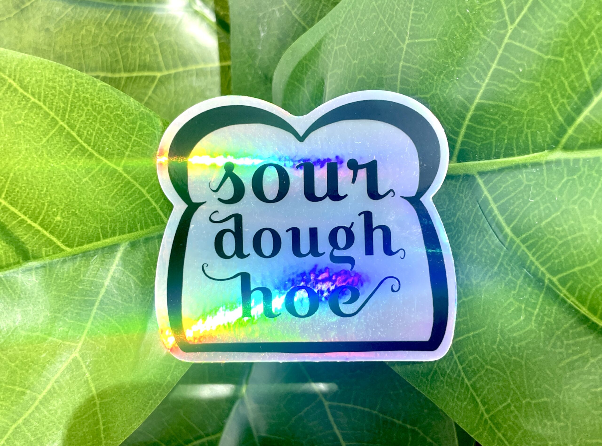 A holographic sticker featuring line art of a slice of bread and the words "sourdough hoe" written in the center.
