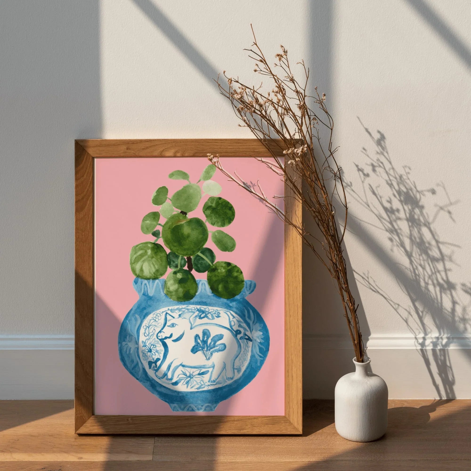 Framed art print of blue vase with green plant, pink background, beside white vase with dried branches