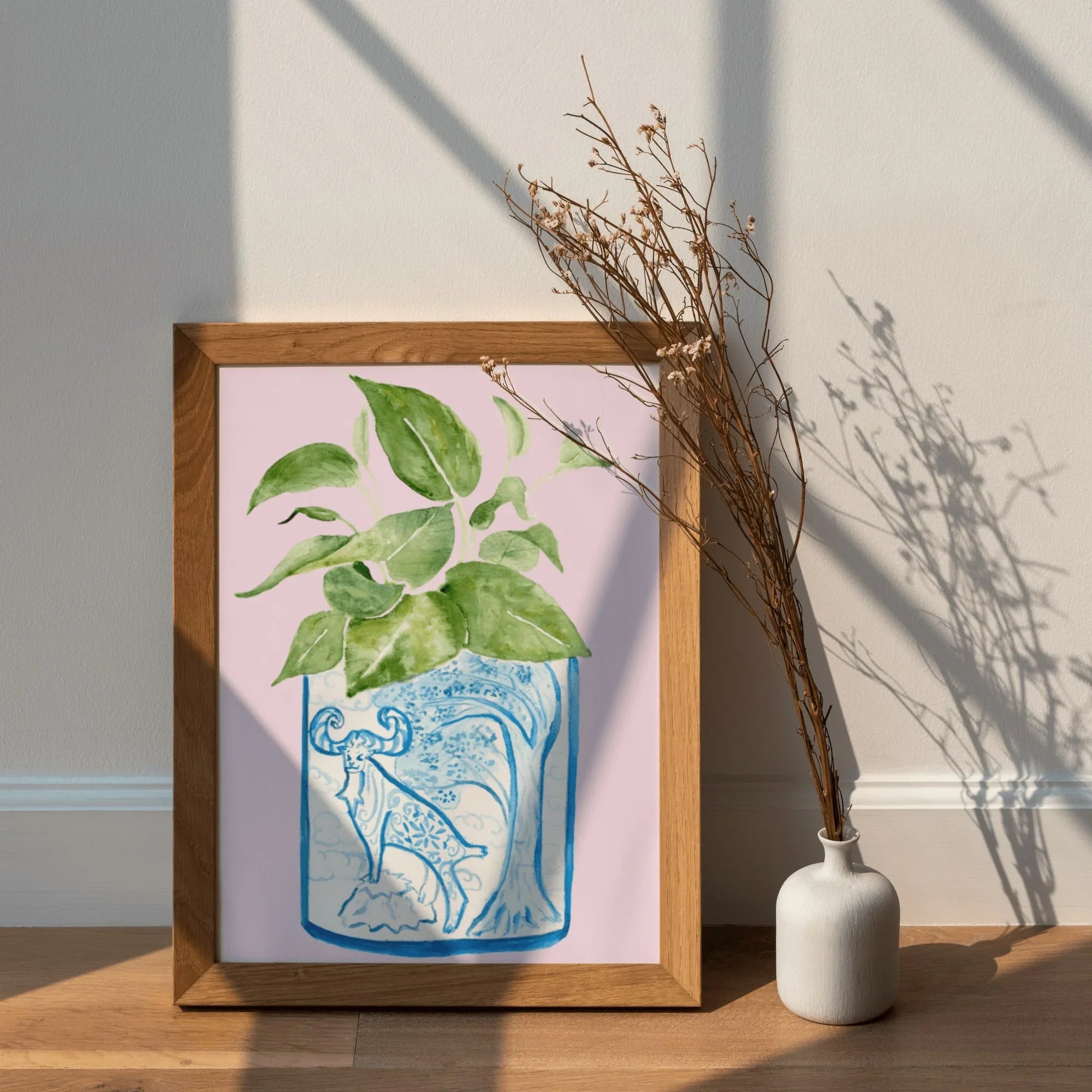 Framed AAPI-inspired art print with green leafy plant in blue patterned vase, styled with dried branches on wood floor