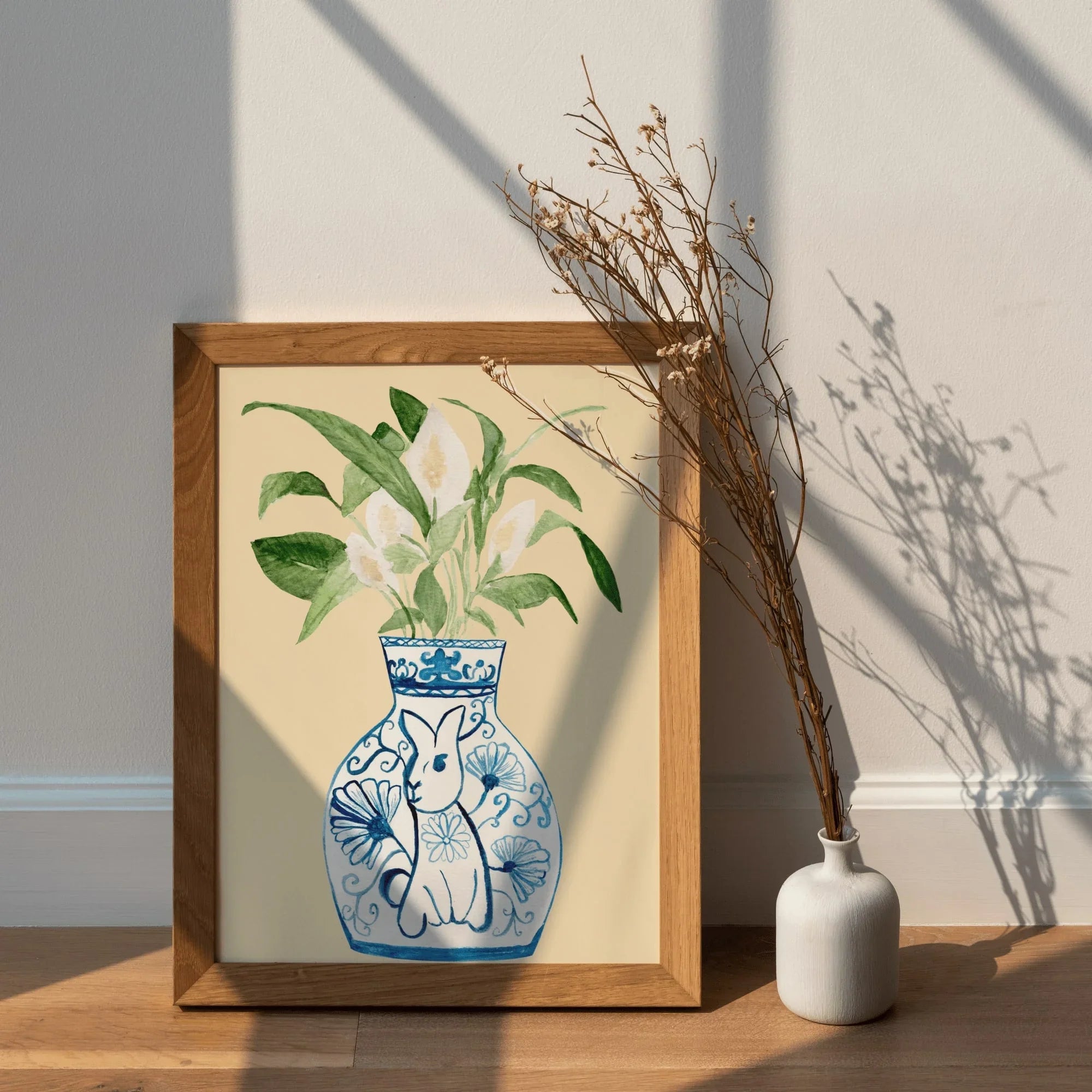 Framed art print of blue and white vase with rabbit and flowers, styled with dried stems on wood shelf