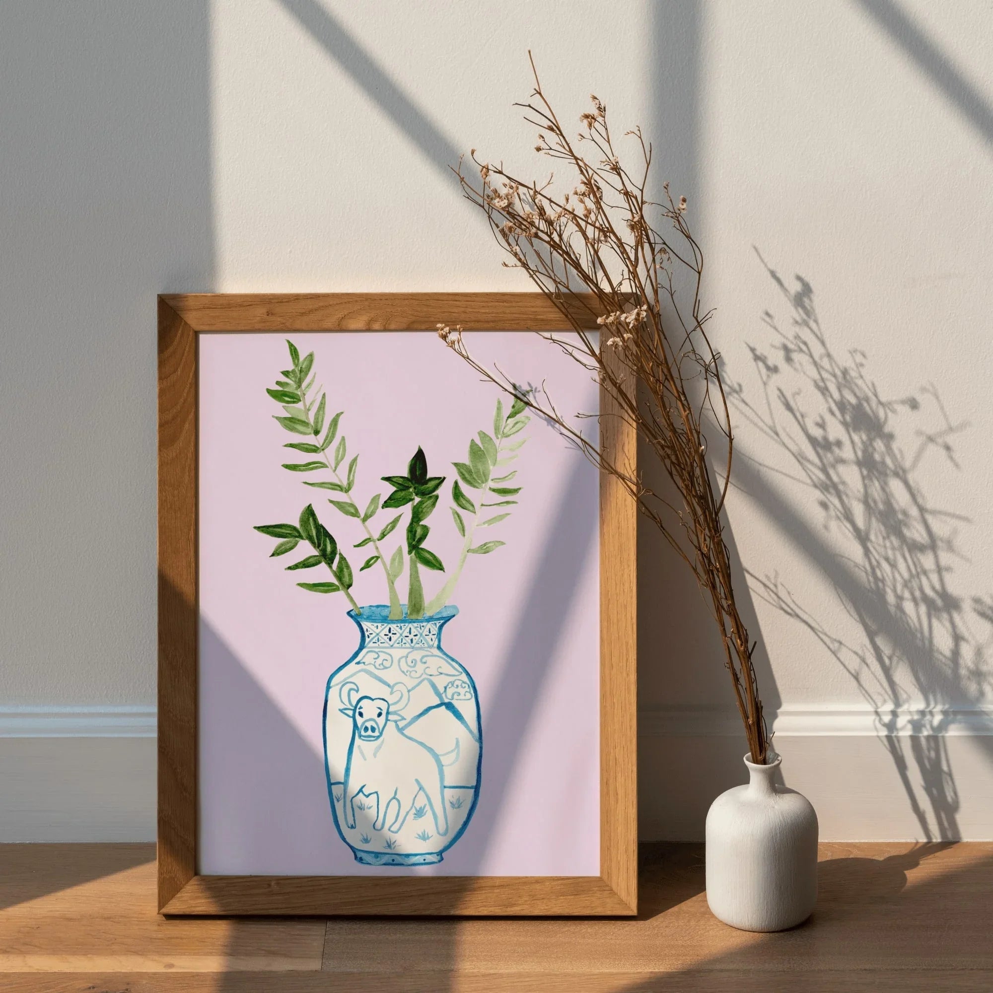 Framed art print of blue and white vase with ox, green leaves, pink background, beside dried flowers in white vase