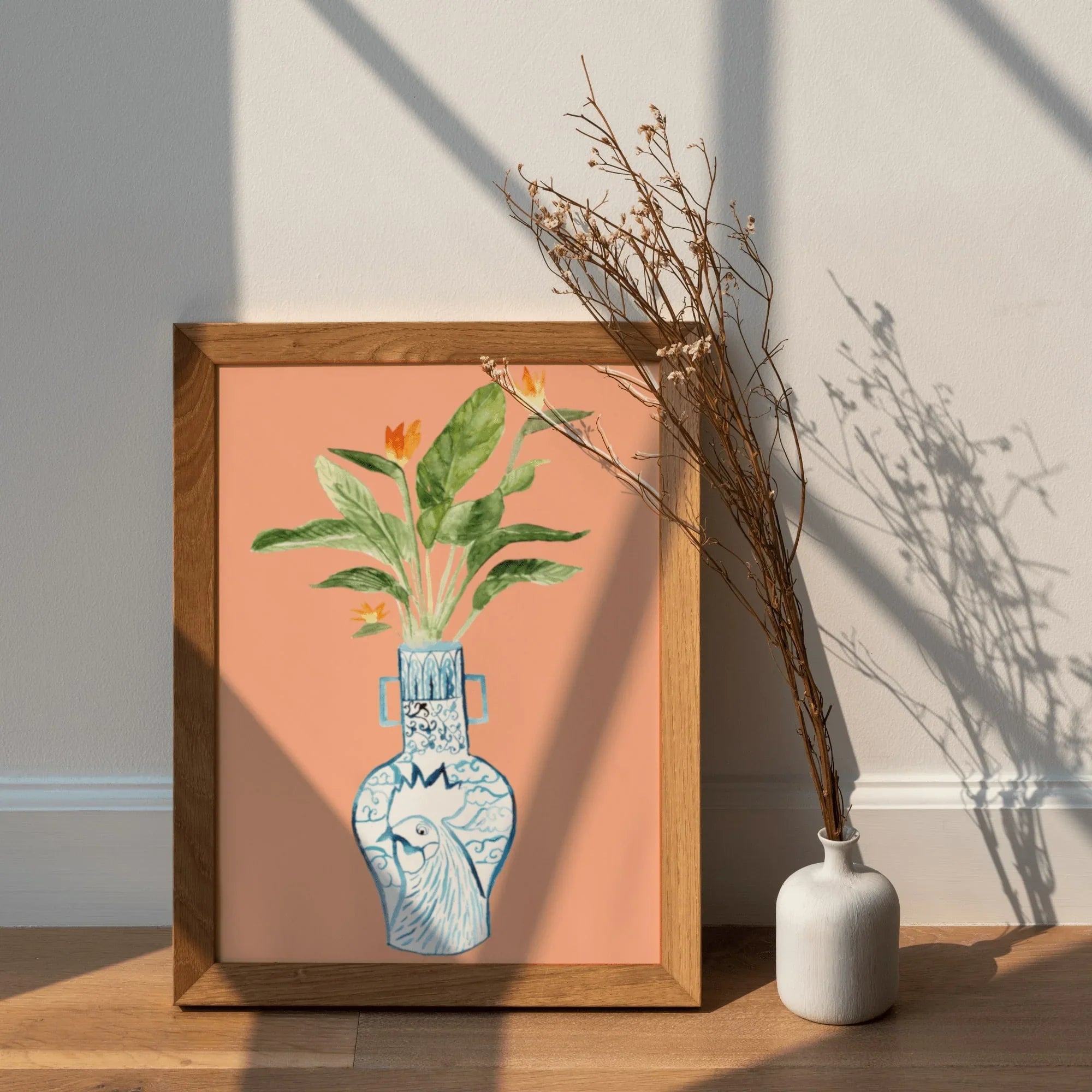 Framed art print of blue patterned vase with bird and green leaves, peach background, beside white vase with dried branches